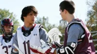 Três jogadores de lacrosse em campo. Dois interagem, um de capacete. O central tem olhar atento.