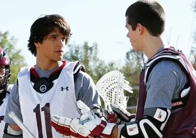 Três jogadores de lacrosse em campo. Dois interagem, um de capacete. O central tem olhar atento.