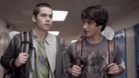 Dois jovens com mochilas em um corredor escolar. Um fala, o outro olha para frente. Um taco de lacrosse é visível.