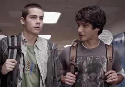 Dois jovens com mochilas em um corredor escolar. Um fala, o outro olha para frente. Um taco de lacrosse é visível.