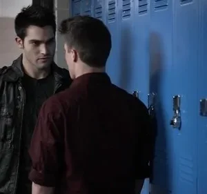 Dois homens num corredor de escola. Um de jaqueta escura encara o outro, de costas, perto de armários azuis. Clima tenso.