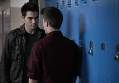 Dois homens num corredor de escola. Um de jaqueta escura encara o outro, de costas, perto de armários azuis. Clima tenso.