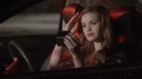 Jovem mulher ruiva ajusta o cabelo, olhando-se no espelho, dentro de um carro escuro com reflexos vermelhos. Chove lá fora, criando um clima tenso.