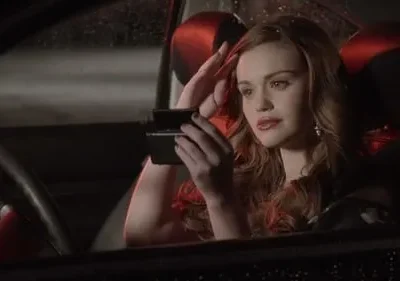 Jovem mulher ruiva ajusta o cabelo, olhando-se no espelho, dentro de um carro escuro com reflexos vermelhos. Chove lá fora, criando um clima tenso.