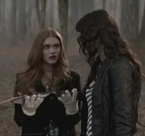 Em floresta nebulosa, mulher ruiva assustada com adaga olha chocada para o lado. Outra mulher de cabelo escuro a observa de perfil.