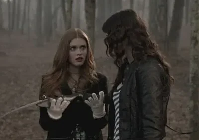 Em floresta nebulosa, mulher ruiva assustada com adaga olha chocada para o lado. Outra mulher de cabelo escuro a observa de perfil.