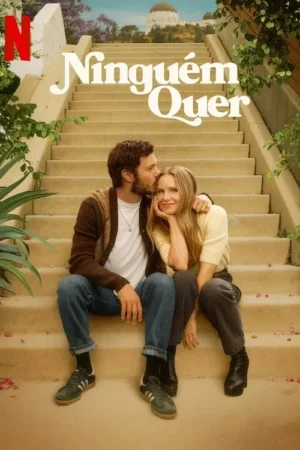 Um casal sorridente sentado em escadas claras. Ele beija a testa dela, que sorri. Ao fundo, um edifício branco com cúpula e céu azul.