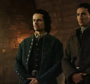 Dois homens sérios em vestes de época numa sala de pedra. Luz natural entra pela janela, criando um ambiente austero e histórico.