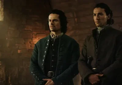 Dois homens sérios em vestes de época numa sala de pedra. Luz natural entra pela janela, criando um ambiente austero e histórico.