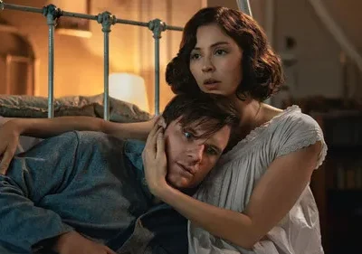 Uma mulher de camisola branca abraça um homem em azul que repousa a cabeça no seu peito. Ambos têm expressões preocupadas em um quarto com iluminação suave.