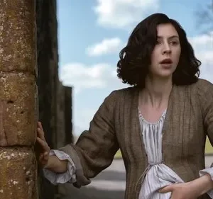 Mulher de época, cabelo escuro, com feição de surpresa e preocupação, toca muro de pedra antigo. Céu azul ao fundo.