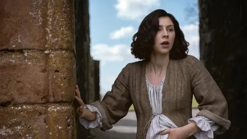 Outlander:Blood of My Blood – T01E06:Direito de Primogenitura 1 Mulher de época,cabelo escuro,com feição de surpresa e preocupação,toca muro de pedra antigo. Céu azul ao fundo.