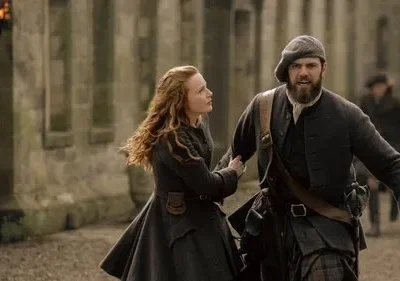 Mulher ruiva e homem barbudo de kilt correm lado a lado em rua de pedra, com castelo ao fundo. Cenário histórico, clima urgente.