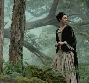 Mulher de vestido de época, verde e creme, olha apreensiva para o lado em uma floresta enevoada com grandes árvores e musgo.