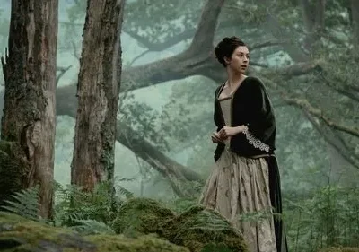 Mulher de vestido de época, verde e creme, olha apreensiva para o lado em uma floresta enevoada com grandes árvores e musgo.