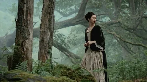 Mulher de vestido de época,verde e creme,olha apreensiva para o lado em uma floresta enevoada com grandes árvores e musgo.