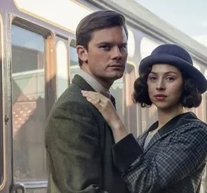 Homem e mulher em trajes de época, com chapéu, olhando fixamente. Ela tem a mão no ombro dele, em frente a um trem antigo.