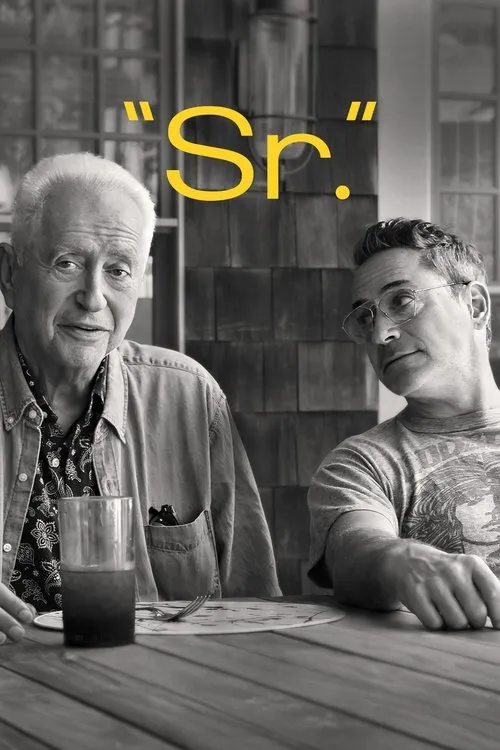 Dois homens em preto e branco à mesa. Um mais velho sorri para a câmera,enquanto um mais jovem com óculos o observa. Cena íntima e reflexiva.