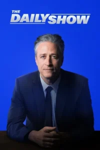 Homem de meia-idade de terno escuro e cabelo grisalho,olhando sério para a frente. Fundo azul vibrante e escrivaninha escura. Mãos unidas.