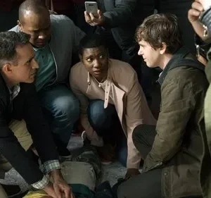 Várias pessoas em torno de alguém no chão. Um homem presta socorro, outro jovem e uma mulher observam com preocupação. Cena de urgência.