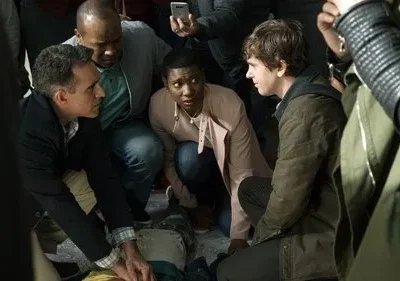 Várias pessoas em torno de alguém no chão. Um homem presta socorro, outro jovem e uma mulher observam com preocupação. Cena de urgência.