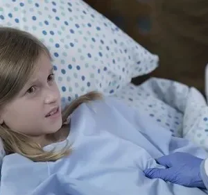 Menina loira em cama hospitalar, com expressão preocupada, enquanto uma mão com luva azul a examina.