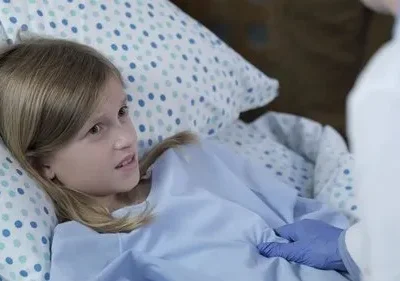 Menina loira em cama hospitalar, com expressão preocupada, enquanto uma mão com luva azul a examina.