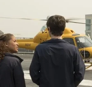 Mulher e homem de uniforme em heliponto, observando helicóptero amarelo. Um piloto aguarda à direita. Cenário urbano sugere missão urgente.