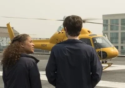 Mulher e homem de uniforme em heliponto, observando helicóptero amarelo. Um piloto aguarda à direita. Cenário urbano sugere missão urgente.