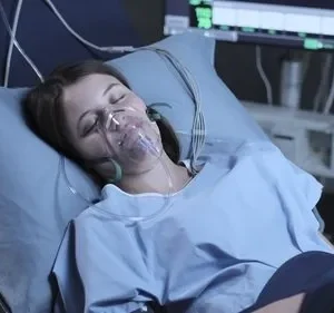 Mulher deitada em leito hospitalar, olhos fechados, com máscara de oxigênio. Monitor de sinais vitais brilhando no escuro ao fundo.