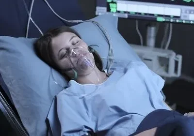 Mulher deitada em leito hospitalar, olhos fechados, com máscara de oxigênio. Monitor de sinais vitais brilhando no escuro ao fundo.