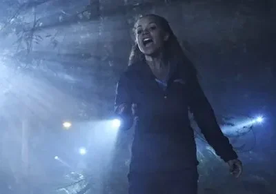 Uma mulher gritando segura uma lanterna em floresta escura e nevoenta. Feixes de luz cortam o ar. Atmosfera de pânico.