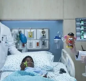 Dois médicos em um quarto de hospital interagem com uma criança doente na cama. Um segura um Sr. Cabeça de Batata para ela. Clima cuidadoso.