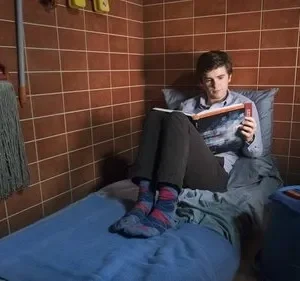Jovem lê um livro numa cama estreita em um quarto minúsculo de azulejos marrons, com vassoura e balde. Clima isolado e calmo.
