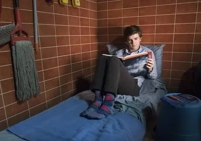 Jovem lê um livro numa cama estreita em um quarto minúsculo de azulejos marrons, com vassoura e balde. Clima isolado e calmo.