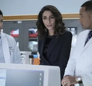 Dois médicos e uma mulher em terno observam um monitor com seriedade em laboratório/hospital.