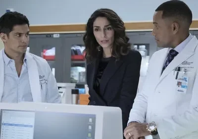 Dois médicos e uma mulher em terno observam um monitor com seriedade em laboratório/hospital.