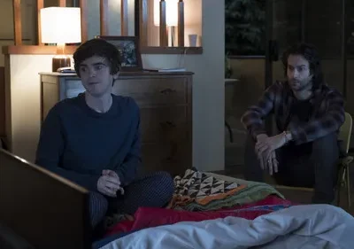 Dois homens num quarto à noite. Jovem de pijama na cama olha atento. Homem barbudo em xadrez senta pensativo numa cadeira. Clima sério.