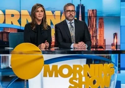 Dois apresentadores sérios em uma bancada de notícias de estúdio, com prédios da cidade ao fundo. Roupas formais.