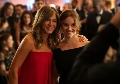 Uma mulher de vestido vermelho e outra de preto sorriem em um evento glamouroso, com convidados ao fundo.