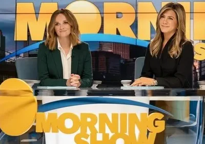 Duas apresentadoras sorridentes sentadas em uma bancada de estúdio de TV. Um painel exibe uma cidade e MORNING SHOW ao fundo.