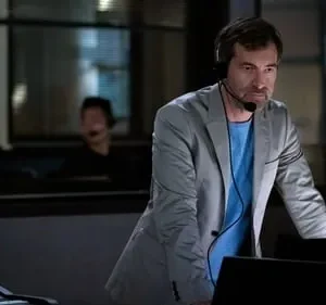 Homem sério de headset e blazer cinza inclina-se sobre um console em ambiente escuro. Pessoas borradas trabalham ao fundo.