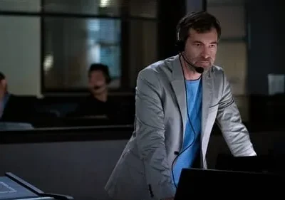 Homem sério de headset e blazer cinza inclina-se sobre um console em ambiente escuro. Pessoas borradas trabalham ao fundo.