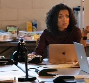 Mulher de cabelo cacheado e suéter roxo olha surpresa em escritório com laptops, papéis e quadro branco.