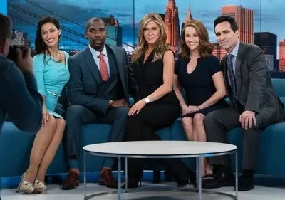 Cinco pessoas posam formalmente em sofá azul num estúdio de TV com skyline. Três mulheres e dois homens, um fotógrafo à esquerda.