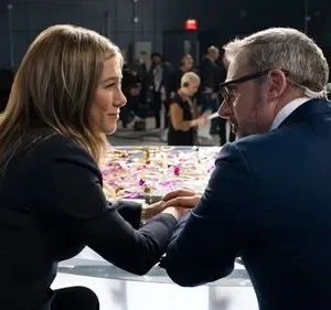 Uma mulher e um homem se encaram em um estúdio de TV, com confetes na mesa. Câmeras e equipe de produção no fundo borrado.