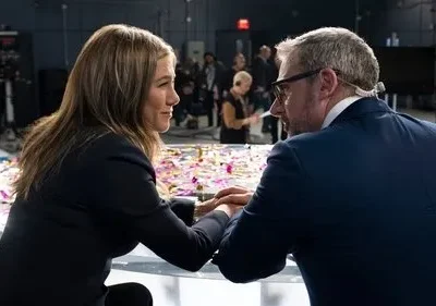 Uma mulher e um homem se encaram em um estúdio de TV, com confetes na mesa. Câmeras e equipe de produção no fundo borrado.