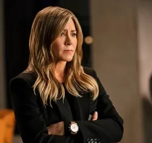 Mulher de blazer escuro e cabelos loiros, braços cruzados, expressão séria, em escritório com globo e cadeira laranja.