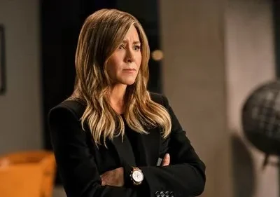 Mulher de blazer escuro e cabelos loiros, braços cruzados, expressão séria, em escritório com globo e cadeira laranja.