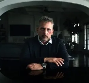 Homem de meia-idade com rosto machucado e expressão séria senta-se à mesa. Fundo de casa elegante, sombrio. Clima dramático.
